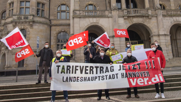Mehrere Personen stehen mit Fahnen und einem Transparent mit der Aufschrift "Tarifvertrag jetzt!!! Landeshauptstadt Hannover ver.di" vor dem hannoverschen Rathaus
